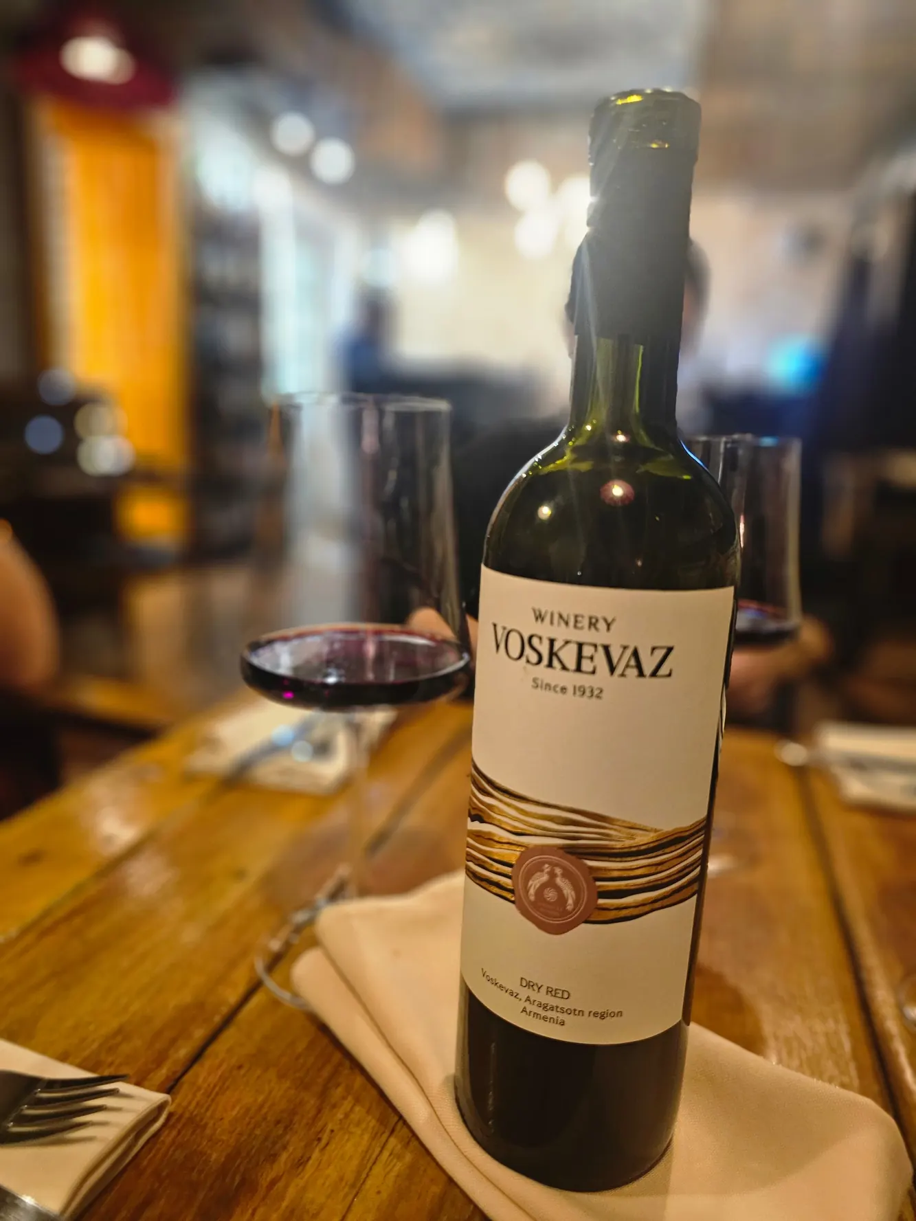 A bottle of Voskevaz red on a wooden table, a glass of red beside it
