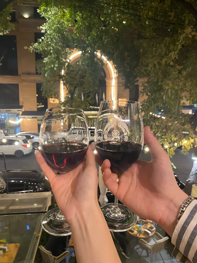 Two glasses of red wine raised outside at night, a lit arch and the trees behind