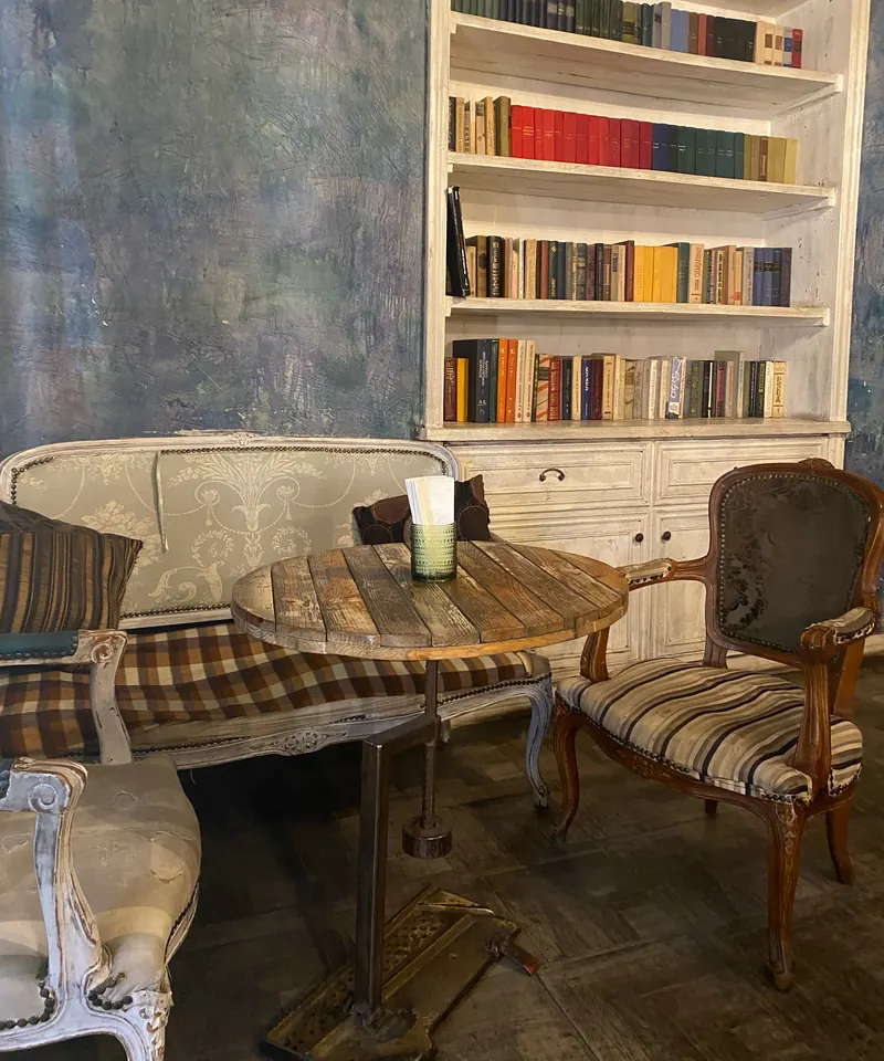 A white bookcase full of old books, a round wooden table and carved armchairs