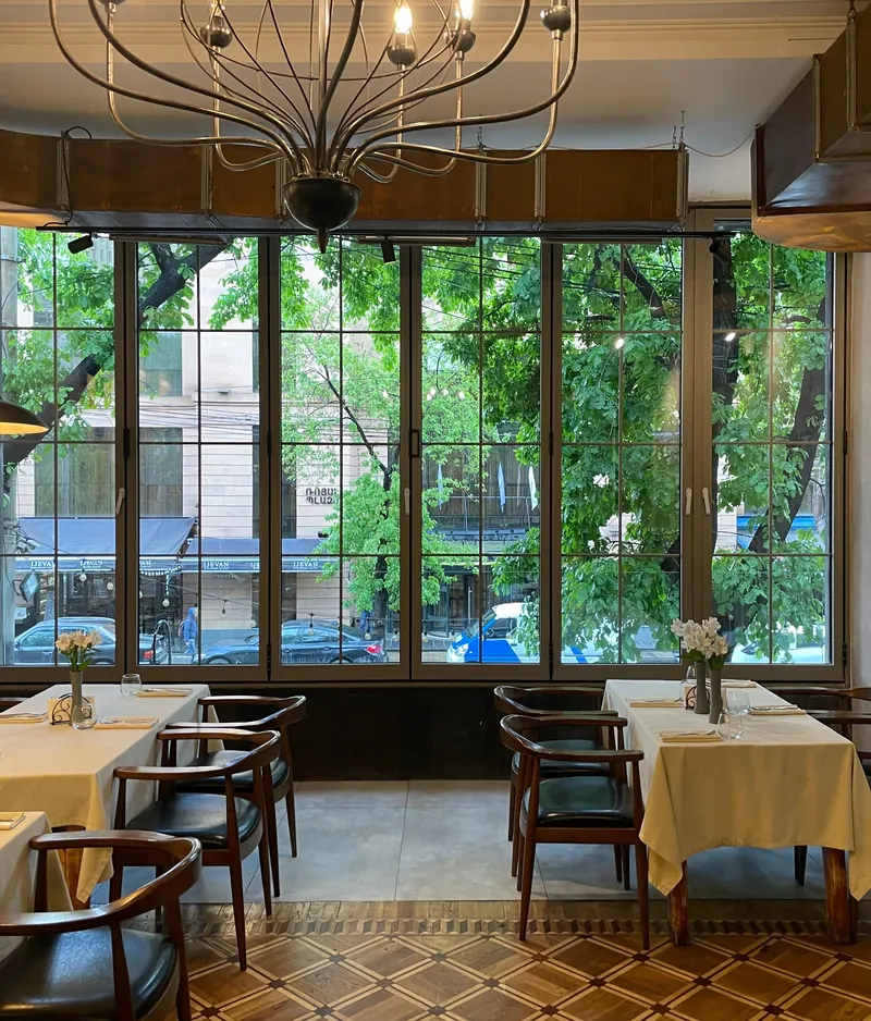 A dining room with yellow tablecloths, wooden armchairs and a tall window onto the trees