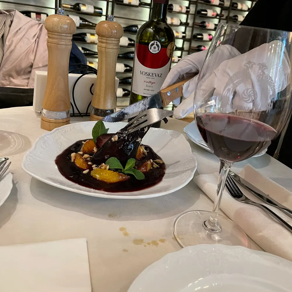 A dish in dark sauce served at a white table, a glass of red and a Voskevaz bottle, the wine wall behind